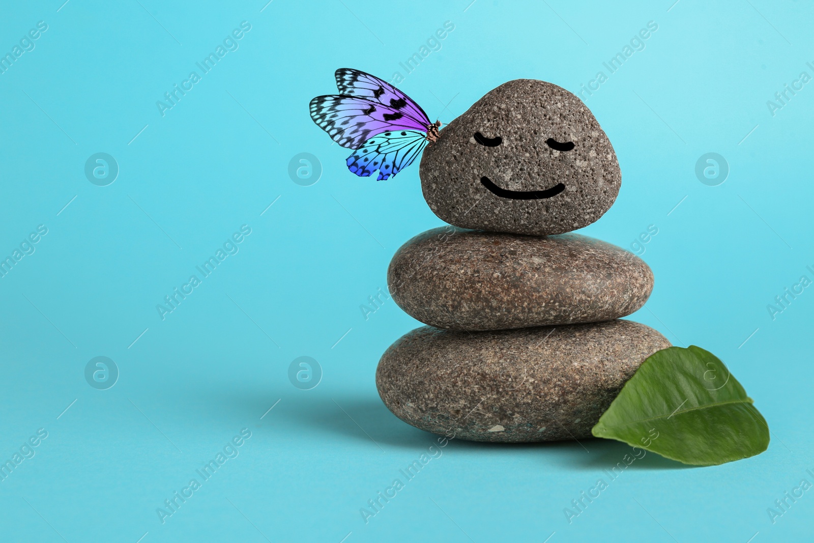 Image of Butterfly on stone with smiling face, stack of pebbles and green leaf on light blue background, space for text. Harmony