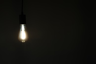 Photo of Glowing light bulb hanging on dark background. Space for text