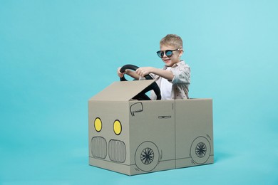 Photo of Little boy in sunglasses driving car made of cardboard on light blue background