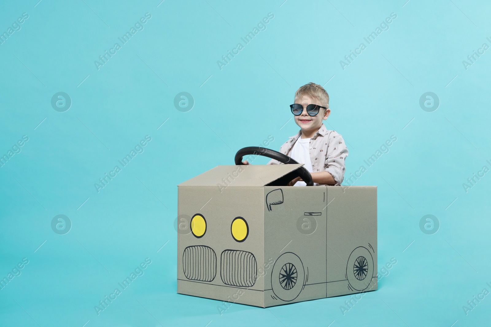 Photo of Little boy in sunglasses driving car made of cardboard on light blue background. Space for text