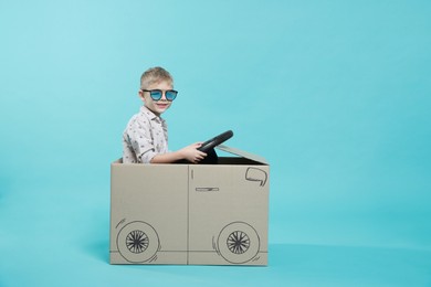 Photo of Little boy in sunglasses driving car made of cardboard on light blue background. Space for text