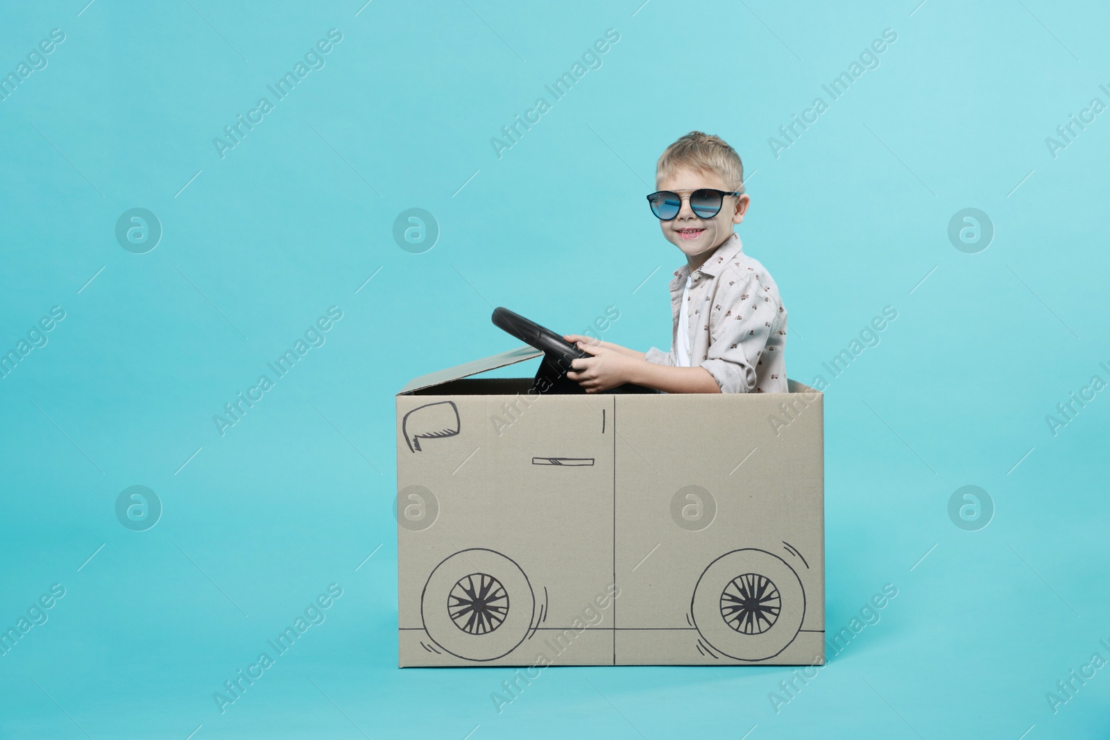 Photo of Little boy in sunglasses driving car made of cardboard on light blue background