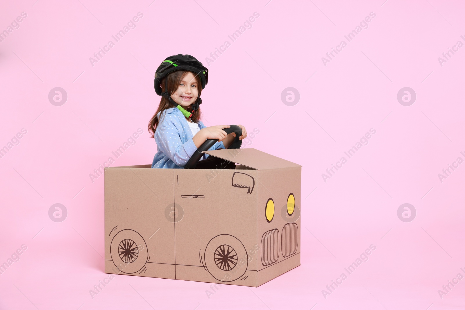 Photo of Little girl driving car made with cardboard on light pink background. Space for text