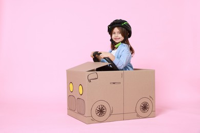 Photo of Little girl driving car made with cardboard on light pink background. Space for text