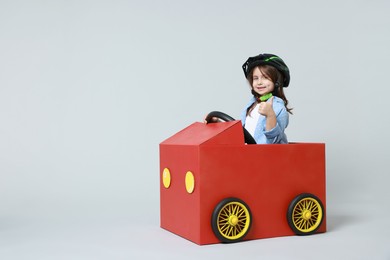 Photo of Little girl showing thumbs up while driving car made with cardboard on light background. Space for text