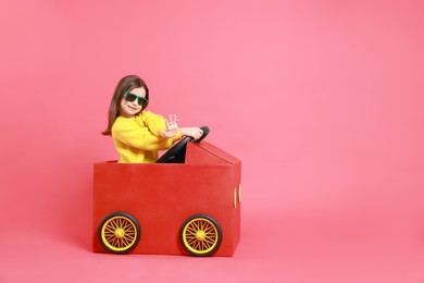 Photo of Little girl in sunglasses driving car made with cardboard on red background. Space for text