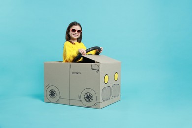 Photo of Little girl in sunglasses driving car made with cardboard on light blue background. Space for text