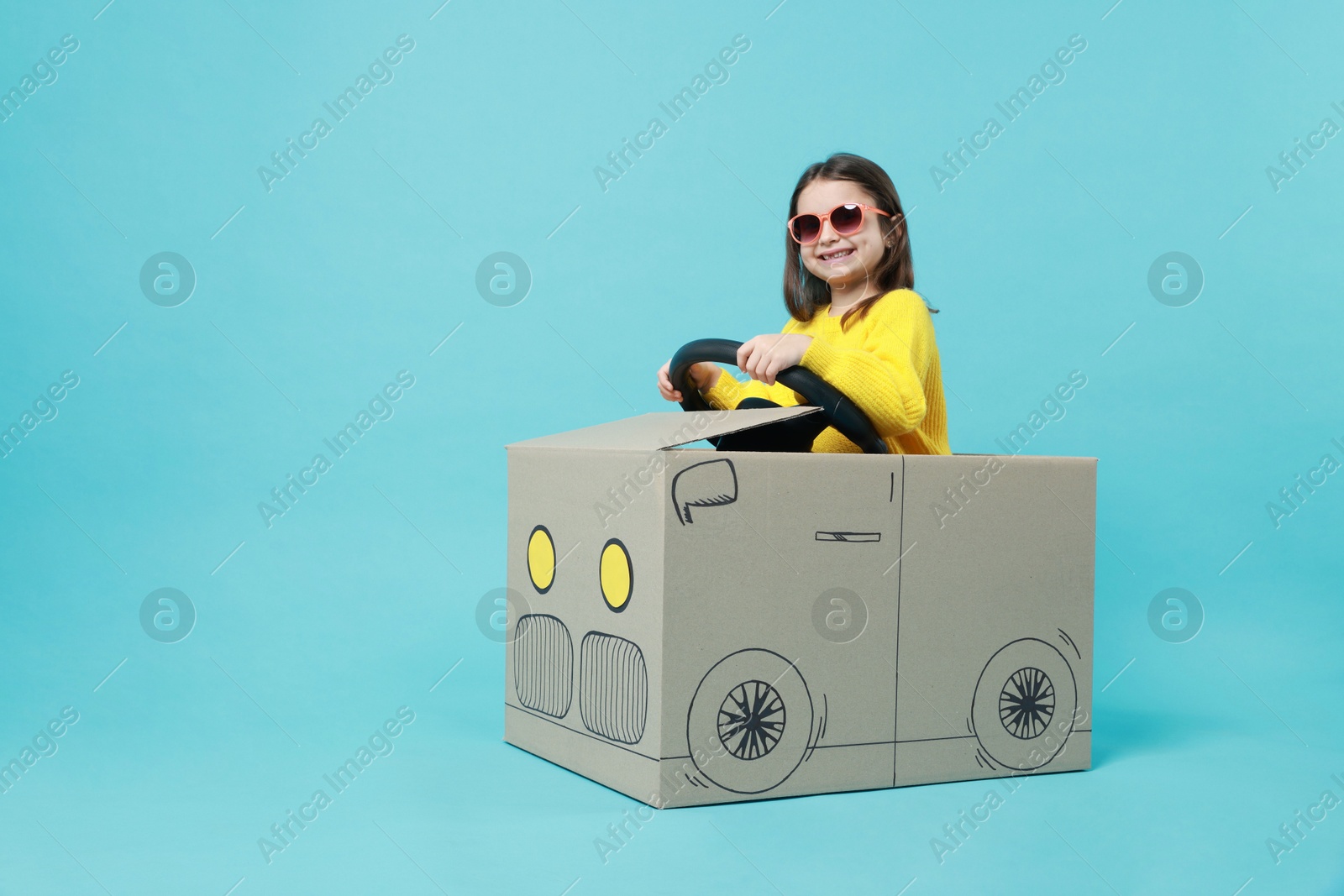 Photo of Little girl in sunglasses driving car made with cardboard on light blue background. Space for text