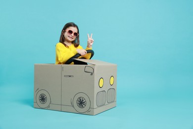 Photo of Little girl showing V-sign while driving car made with cardboard on light blue background. Space for text