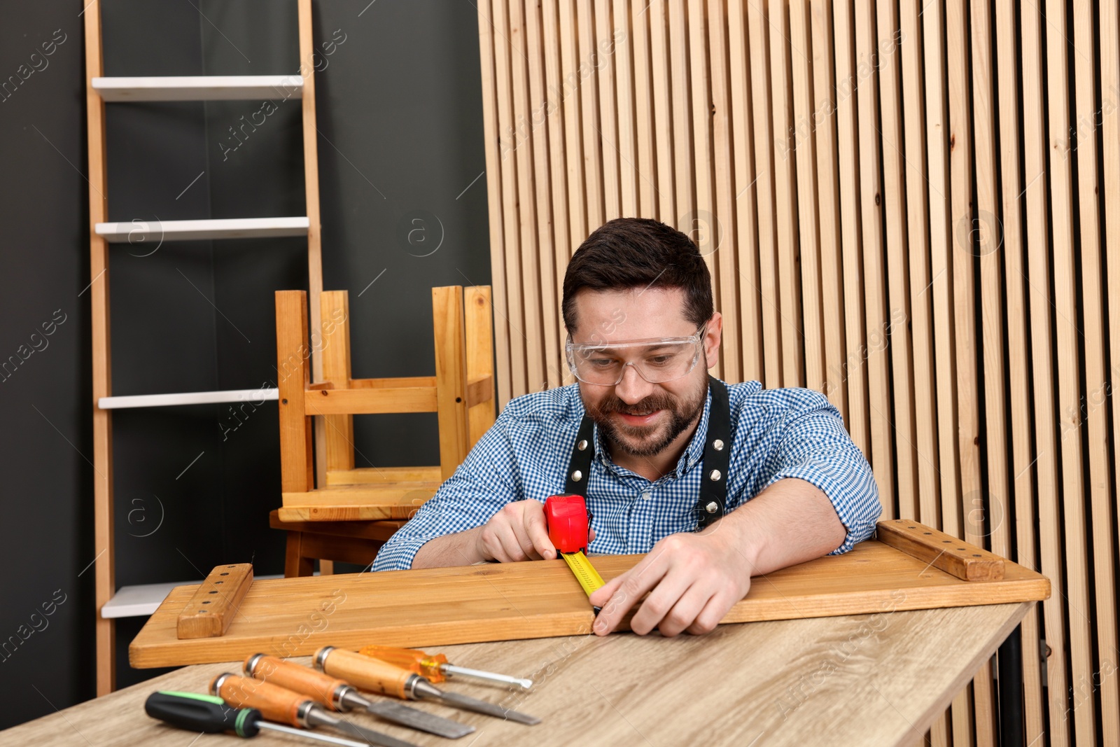 Photo of Relaxing hobby. Smiling man measuring wooden plank with tape at table in workshop