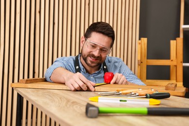 Photo of Relaxing hobby. Smiling man measuring wooden plank with tape at table in workshop