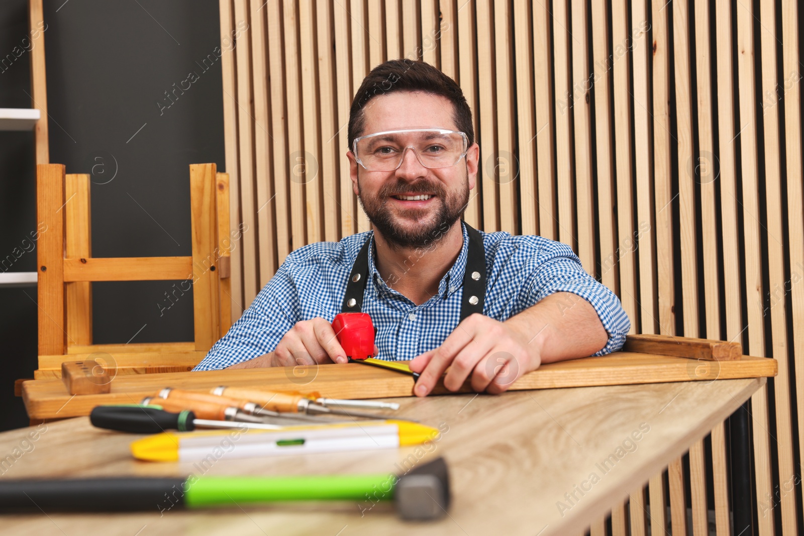 Photo of Relaxing hobby. Smiling man measuring wooden plank with tape at table in workshop