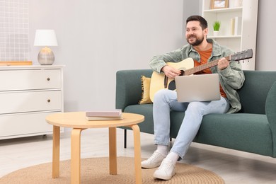 Photo of Relaxing hobby. Smiling man playing guitar with online tutorial by laptop on sofa at home