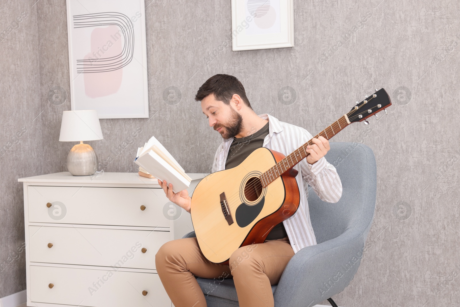 Photo of Relaxing hobby. Man with guitar and book on armchair at home