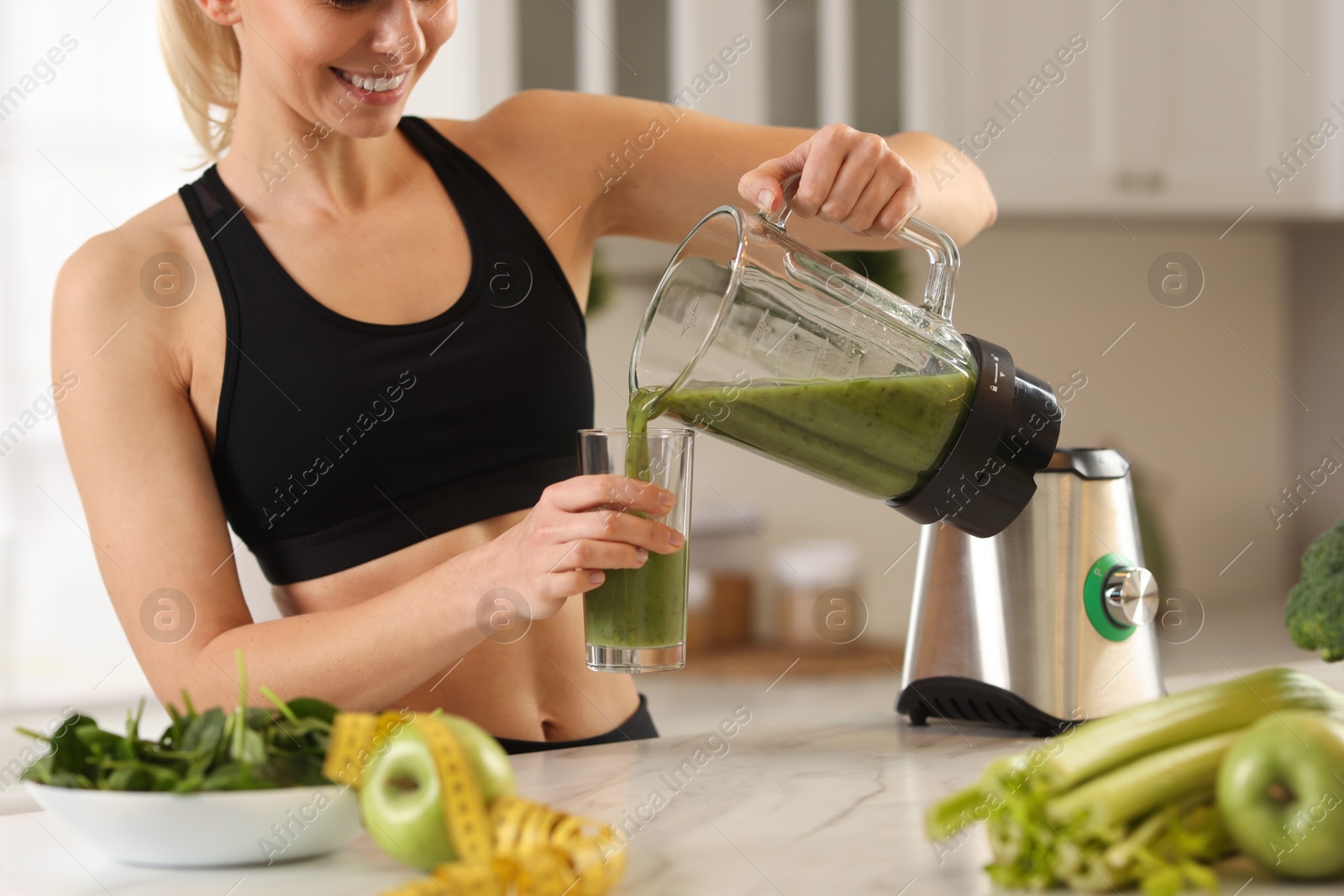 Photo of Weight loss. Woman pouring fresh shake from blender cup into glass at white marble table in kitchen, closeup