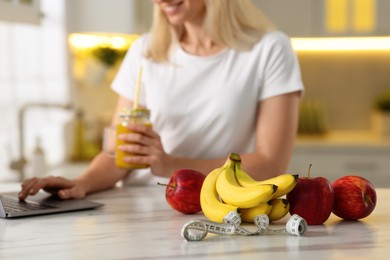 Photo of Weight loss. Woman with tasty shake using laptop at white marble table in kitchen, focus on fresh fruits and measuring tape