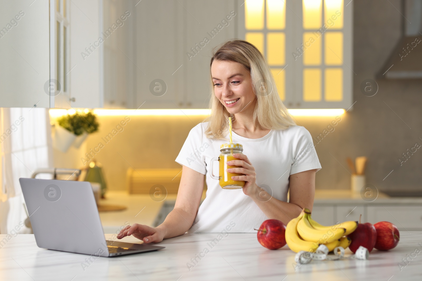 Photo of Weight loss. Happy woman with tasty shake using laptop at white marble table in kitchen