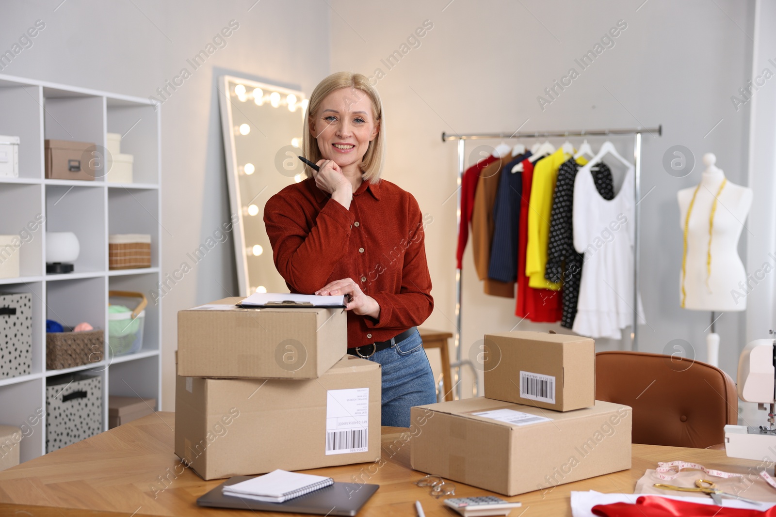 Photo of Business owner with clipboard and pen near parcels in her tailor shop