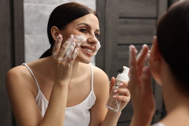 Photo of Woman washing her face with cleansing foam near mirror in bathroom