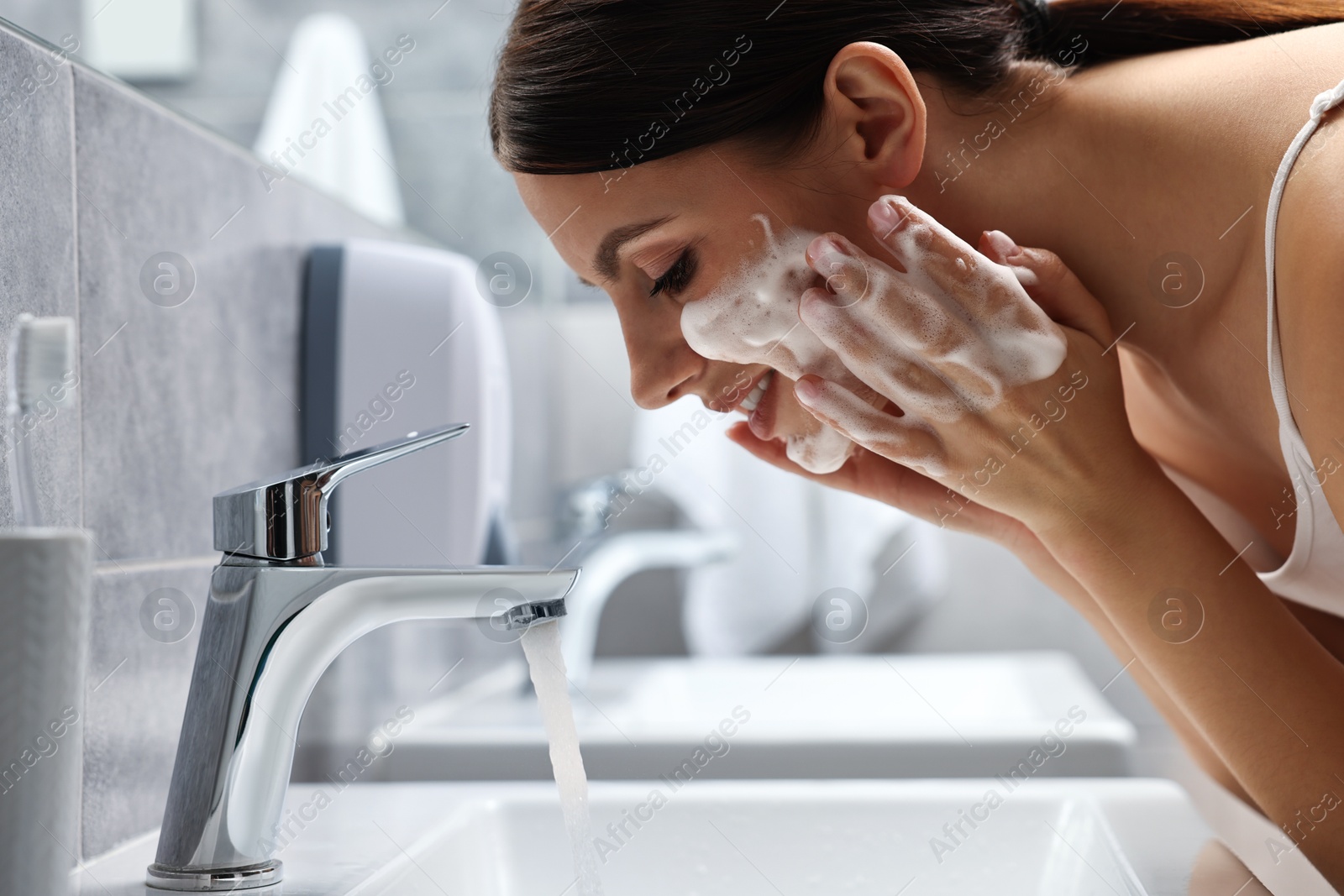 Photo of Woman washing her face with cleansing foam over sink in bathroom