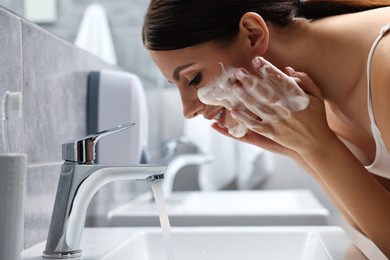 Photo of Woman washing her face with cleansing foam over sink in bathroom