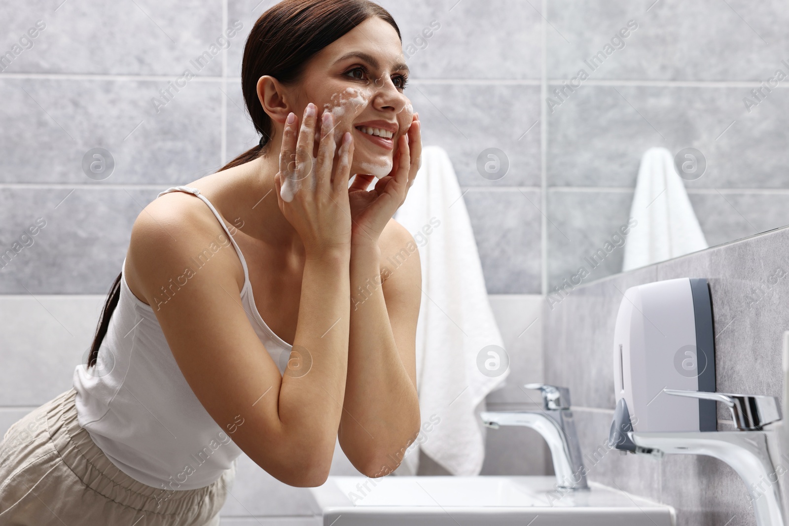 Photo of Woman washing her face with cleansing foam in bathroom