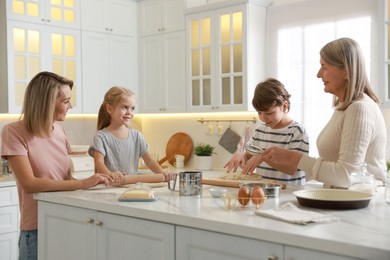 Photo of Little kids with their mother and grandmother making dough at white marble table in kitchen