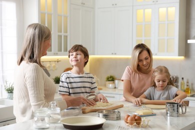 Photo of Little kids with their mother and grandmother making dough at white marble table in kitchen