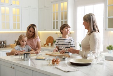 Photo of Little kids with their mother and grandmother making dough at white marble table in kitchen