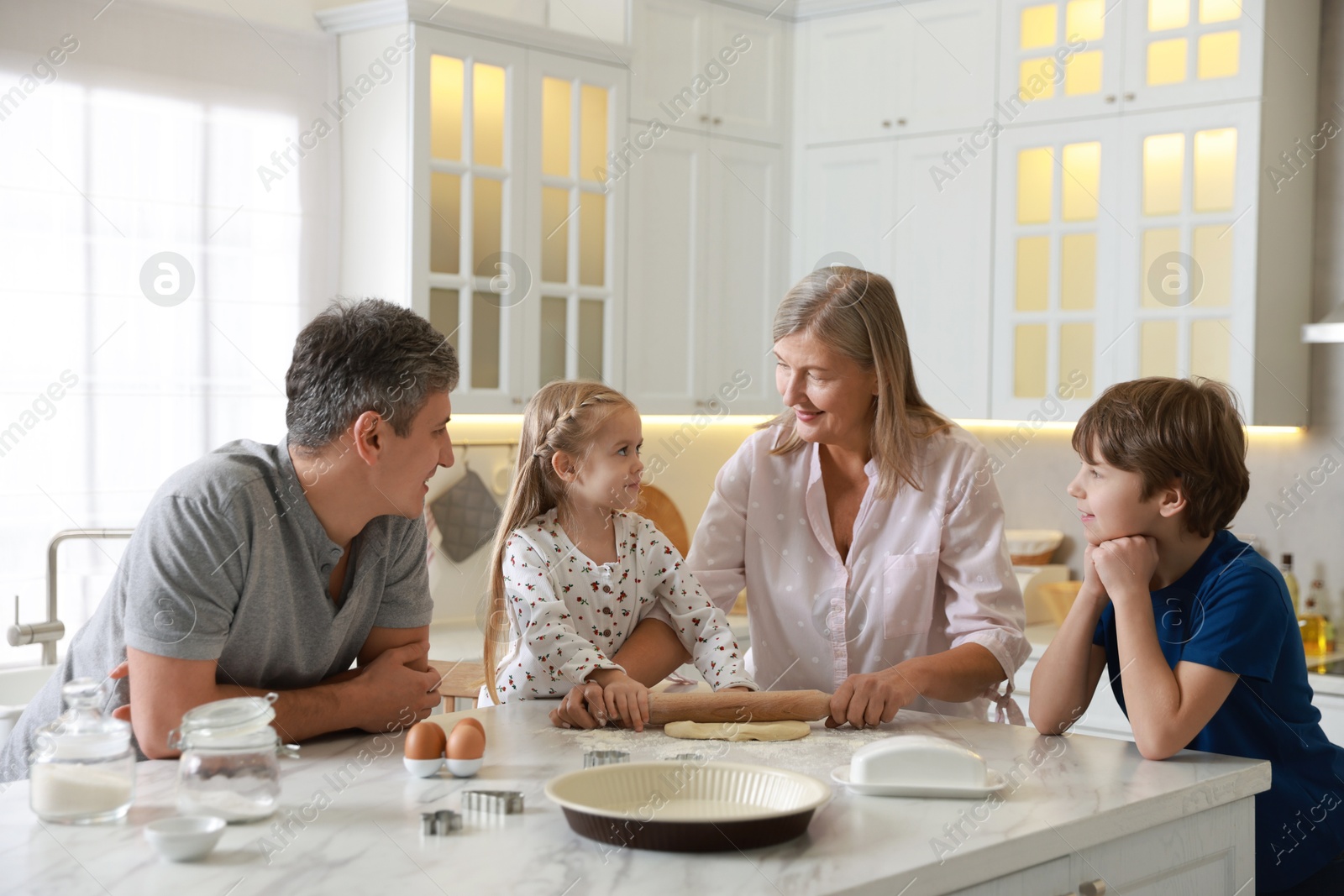 Photo of Little kids with their father and grandmother making dough at marble table in kitchen