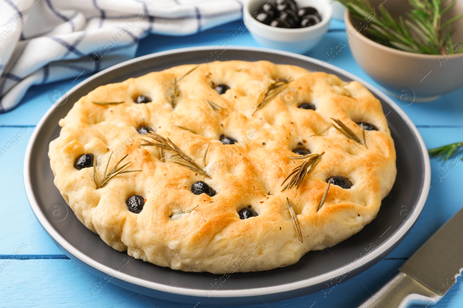 Delicious focaccia bread with olives, rosemary and knife on light blue wooden table, closeup Photo of Delicious focaccia bread with olives, rosemary and knife on light blue wooden table, closeup