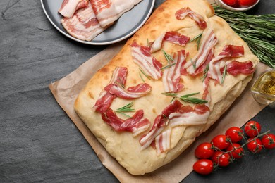 Photo of Delicious focaccia bread with bacon, rosemary, oil and tomatoes on black table, flat lay