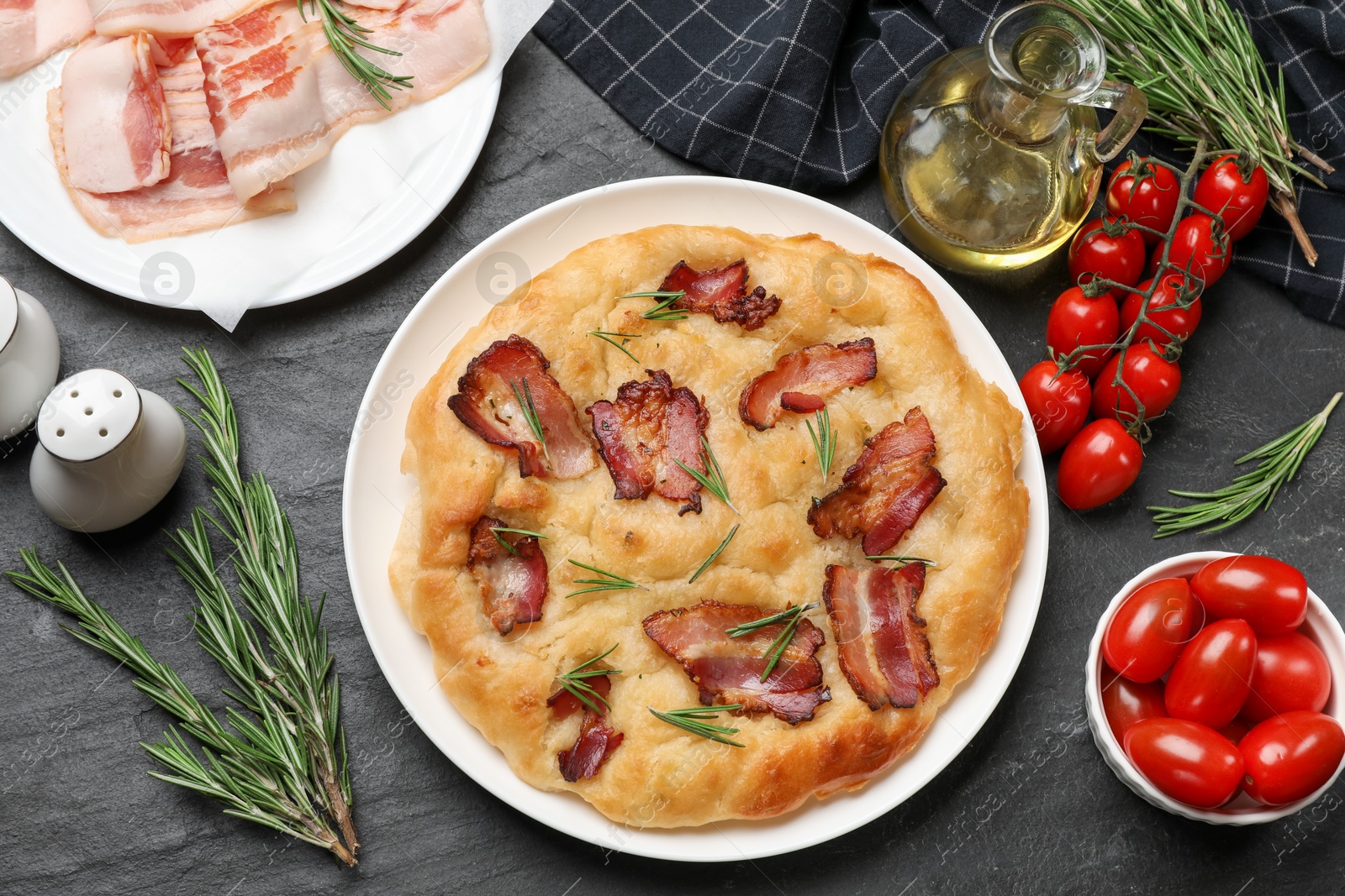 Photo of Delicious focaccia bread with bacon, rosemary, oil and tomatoes on black table, flat lay