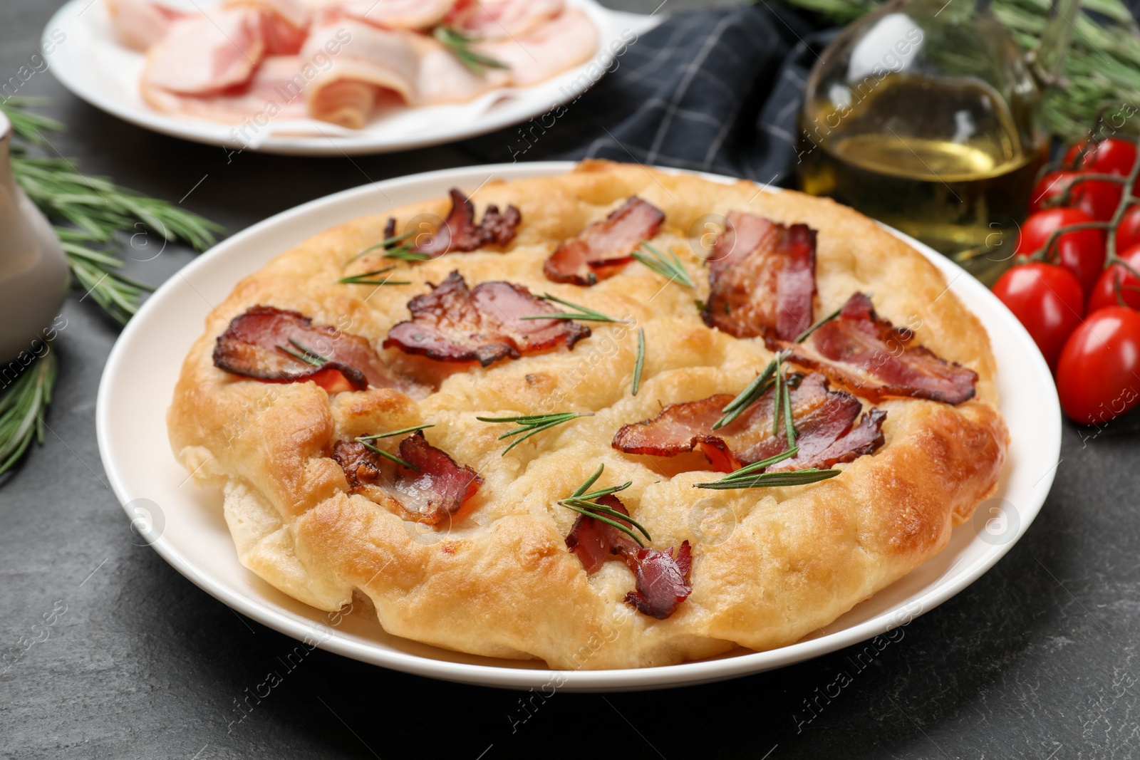 Photo of Delicious focaccia bread with bacon, rosemary, oil and tomatoes on black table, closeup