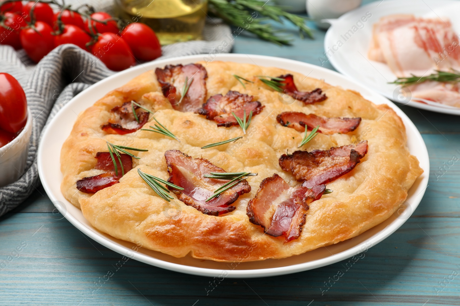 Photo of Delicious focaccia bread with bacon, rosemary and tomatoes on light blue wooden table, closeup