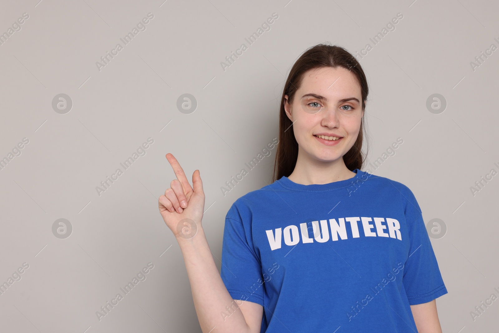 Photo of Portrait of smiling woman in t-shirt with word Volunteer pointing at something on grey background. Space for text