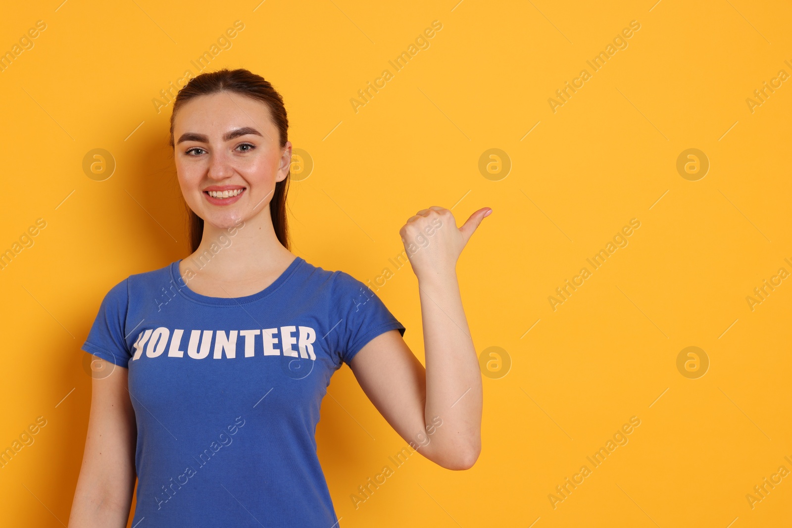 Photo of Portrait of smiling woman in t-shirt with word Volunteer pointing at something on orange background. Space for text