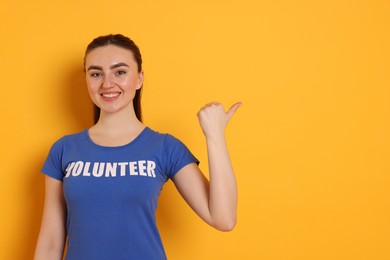 Photo of Portrait of smiling woman in t-shirt with word Volunteer pointing at something on orange background. Space for text