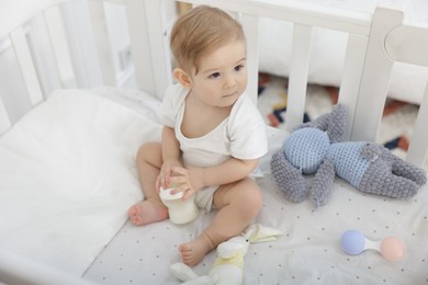 Photo of Cute little baby with feeding bottle of milk in crib at home