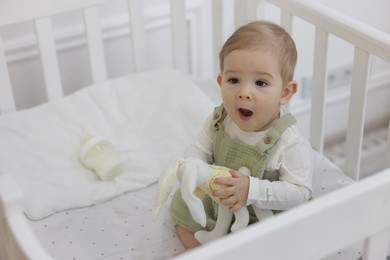 Photo of Cute little baby with toy bunny and feeding bottle of milk in crib at home, space for text