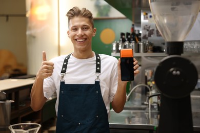 Photo of Smiling cafe worker with payment terminal showing thumbs up indoors