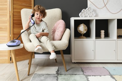 Photo of Cute little boy with telephone in armchair indoors