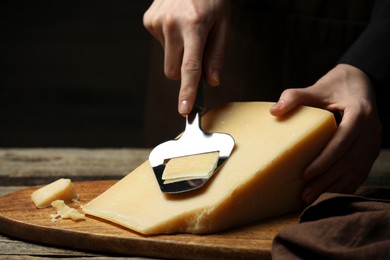 Photo of Woman cutting delicious cheese with slicer at wooden table against black background, closeup