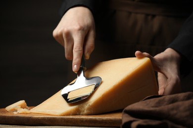 Photo of Woman cutting delicious cheese with slicer at wooden table against black background, closeup