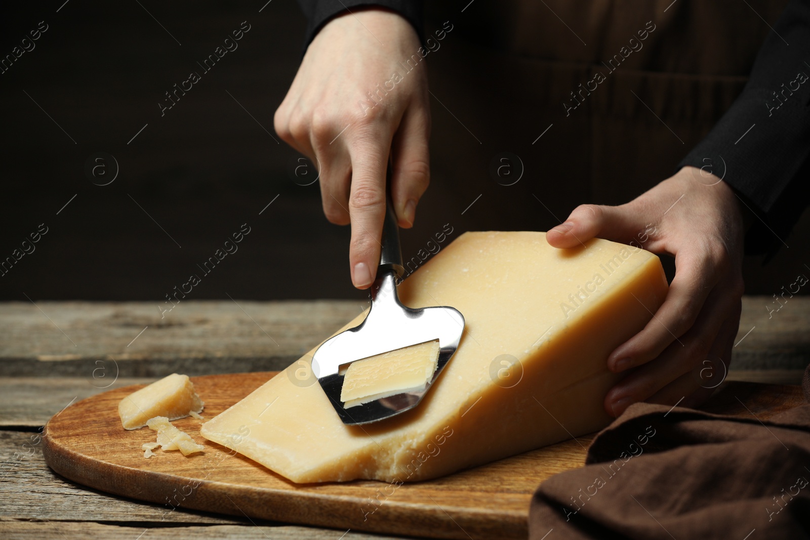 Photo of Woman cutting delicious cheese with slicer at wooden table against black background, closeup