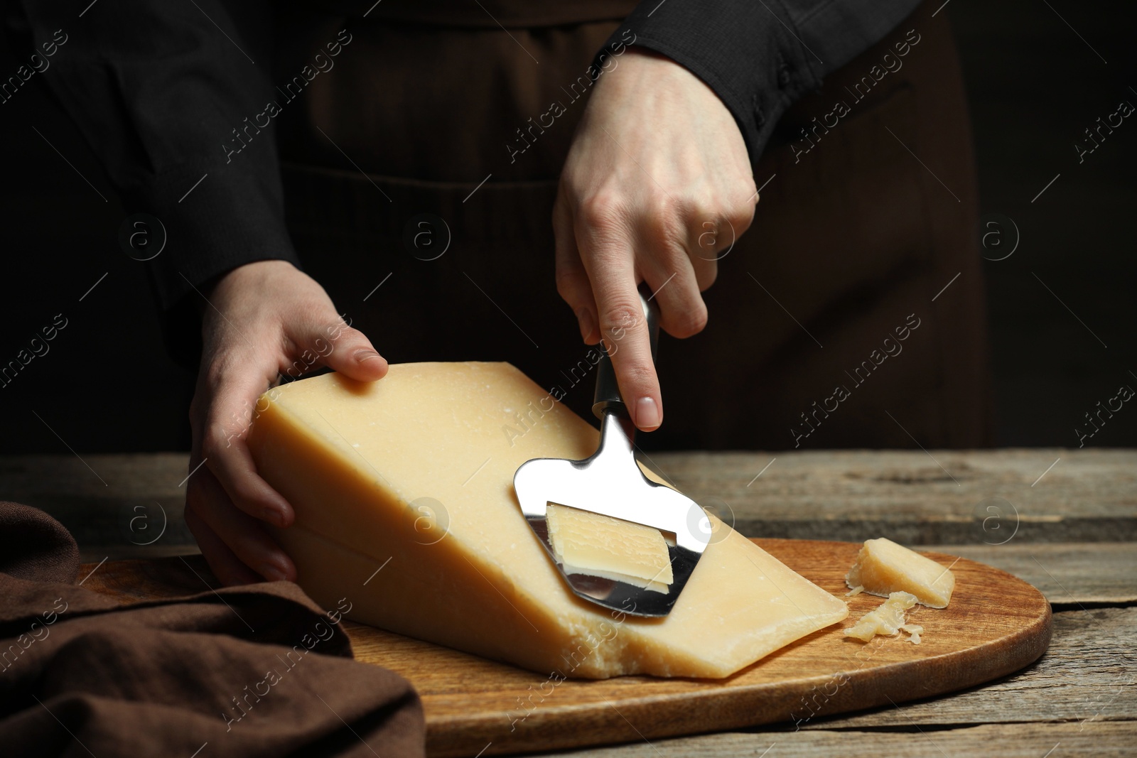 Photo of Woman cutting delicious cheese with slicer at wooden table against black background, closeup