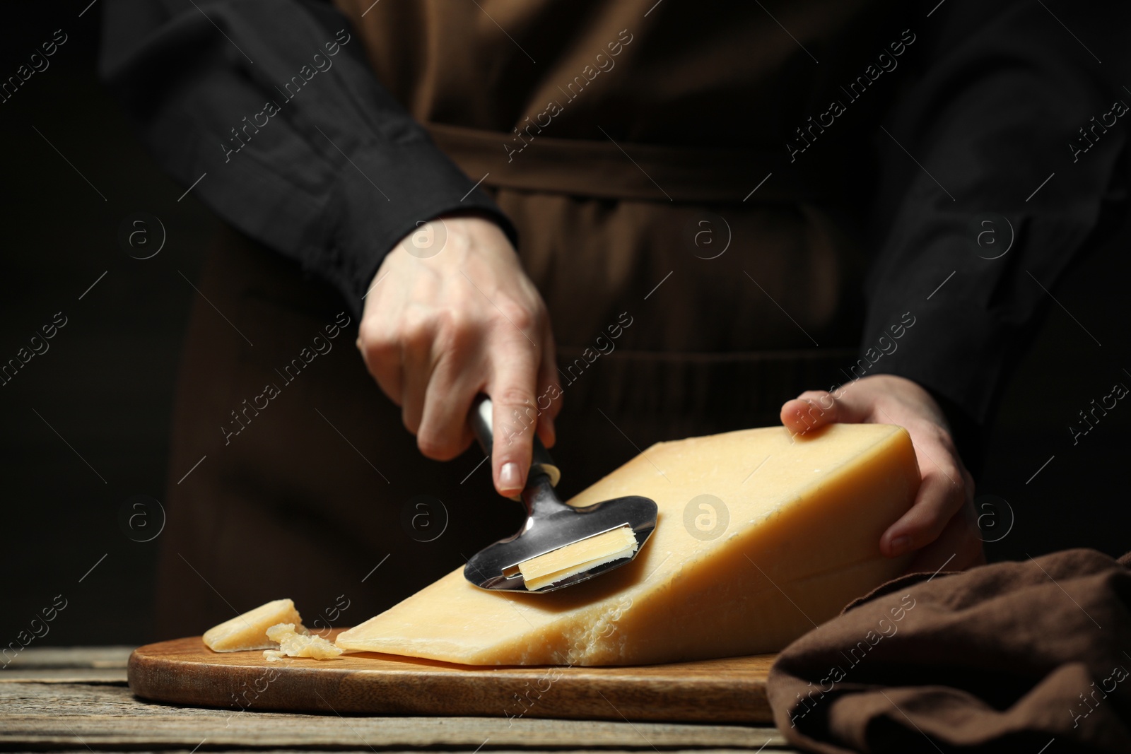 Photo of Woman cutting delicious cheese with slicer at wooden table against black background, closeup