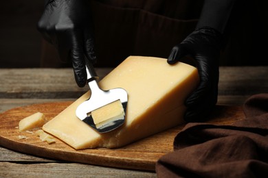 Photo of Woman in gloves cutting delicious cheese with slicer at wooden table against black background, closeup