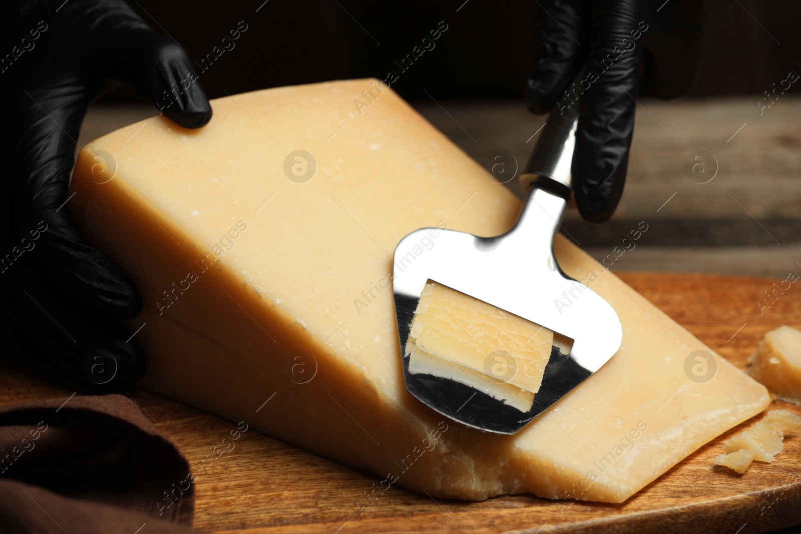 Photo of Woman in gloves cutting delicious cheese with slicer at wooden table against black background, closeup