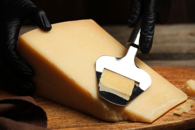 Photo of Woman in gloves cutting delicious cheese with slicer at wooden table against black background, closeup